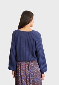 Lila gerippter Pullover mit Ballonärmel, der einen weiten Halsausschnitt und eine weiche Textur aufweist. Kombiniert mit einem gemusterten Rock in Blau- und Weinrot-Tönen.