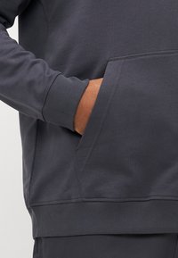 Man's hand inside front pocket of dark gray sweatshirt with ribbed cuff and hem, visible from waist to mid-torso.