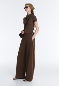 Femme portant un pantalon marron à jambes larges et un polo, foulard vert sur la tête, lunettes de soleil noires et sac en bandoulière, debout avec une main dans la poche.