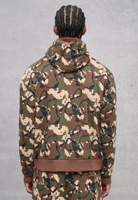 Persona con capelli intrecciati che indossa un hoodie a motivo camouflage e pantaloni abbinati, in piedi di fronte a un muro grigio uniforme, rivolto all'indietro.