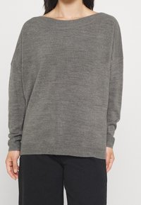 Grau gestrickter Pullover mit weitem Bootsausschnitt, langen Ärmeln und gerippten Bündchen. Weiche Textur und lässige Passform, kombiniert mit schwarzen Hosen.