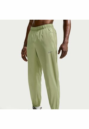 Homme portant un pantalon de jogging Nike vert clair avec taille élastique et chevilles élastiquées, debout avec les bras détendus le long du corps sur un fond uni.
