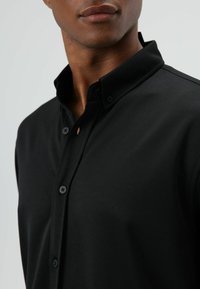 Zwart overhemd met knoopsluiting, een gestructureerde kraag, een gladde textuur en knopen aan de voorkant. Een opvallend detail is een kleine opening vlak bij de kraag.