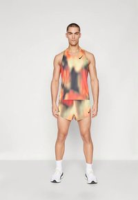 Sportlicher Mann steht mit dem Gesicht nach vorn, trägt ein orangefarbenes, schwarzes und gelbes Batik-Ärmelshirt sowie passende Shorts, dazu weiße Sneaker und Socken.