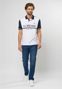 Hvid polo-shirt med marinefarvede ærmer, der har et horisontalt logodesign. Bæres med blå jeans og hvide sneakers, stående på en ensfarvet baggrund.