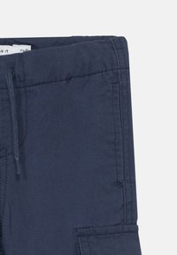 Námornícke modré nohavice vyrobené z ľahkého materiálu s elastickým pásom, bočnými vreckami a rovnými nohavicami. Viditeľná jemná textúra.