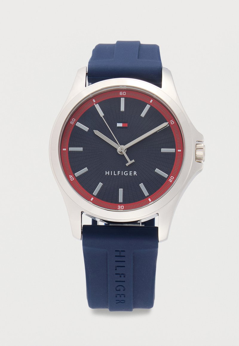 Reloj de pulsera para hombre con esfera azul, anillo de minutos rojo, marcadores y agujas plateados, correa de silicona azul grabada con "Hilfiger" y caja plateada.