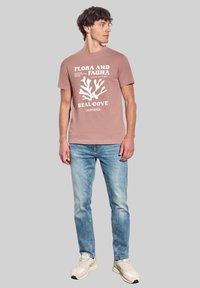 Bomullströja i dammig rosa med ett vitt koralldesign och texten "Seal Cove California", kombinerad med ljusblå jeans och sneakers.