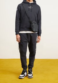 Mörkgrå hoodie med metallisk logotyp, matchande joggers med sidofickor, svartvita sneakers och en liten svart axelremsväska. Gul bakgrund.