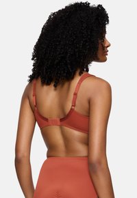 Ženska s kodrastimi lasmi, oblečena v rjavo-braon modrček in hlačke s visokim pasom, prikazana s hrbtne strani na enobarvni svetli podlagi.