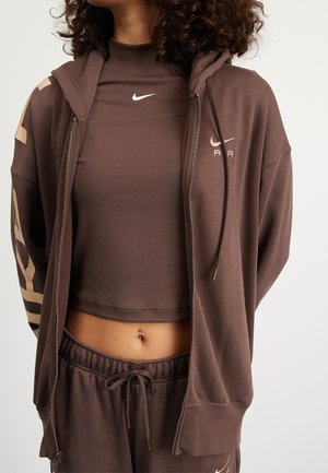 Femme portant un sweat à capuche zippé Nike marron, un col roulé court assorti et un pantalon de survêtement à cordon, montrant un piercing au nombril.
