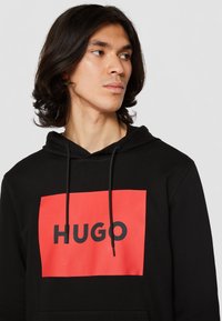 Černá mikina s kapucí a červeným čtvercem, na kterém je v tučném černém písmu napsáno "HUGO". Má stahovací kapuci a příjemný jemný materiál.