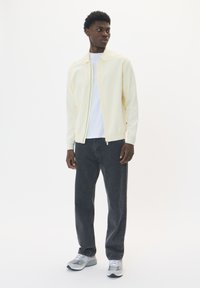 Jachetă crem cu fermoar peste o cămașă albă, asortată cu blugi gri închis și pantofi sport gri. Material neted, croială modernă, design simplu.