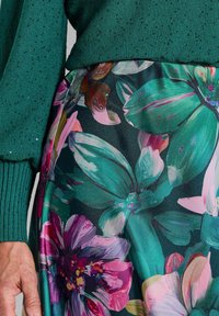 Pull en tricot vert avec des poignets côtelés, associé à une jupe fleurie ornée de fleurs vertes et roses vives sur un fond sombre.