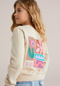 Meitene ar garām blondām matiem, tērpusies bēša džemperī ar krāsainiem abstraktiem ziedu rakstiem aizmugurē, kopā ar gaiši ziliem džinsiem.