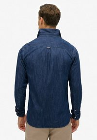 Donkerblauwe denim blouse met lange mouwen, knopenmanchetten en een hoge kraag. Heeft een achternaaddetail en een lichte textuur.