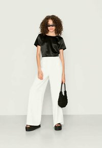 Mulher com cabelo encaracolado, vestindo uma blusa de cetim preta, calças largas brancas, sandálias de plataforma pretas, óculos de sol, segurando uma bolsa texturizada preta.