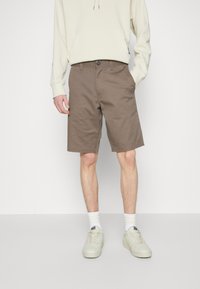Shorts en coton marron à coupe droite, avec poches latérales et fermeture à bouton, associés à un sweat à capuche léger et des baskets blanches.