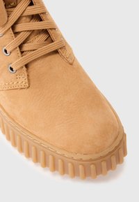 Bottine en daim beige avec semelle en caoutchouc épaisse et crantée, ainsi que des lacets tressés assortis passés à travers des œillets en métal sur fond blanc.