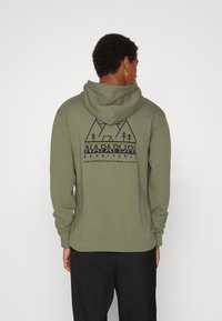 Sweat à capuche vert olive avec une capuche doublée. Présente un imprimé graphique de montagnes et d'arbres en bleu marine au dos. Tissu doux avec des poignets côtelés.