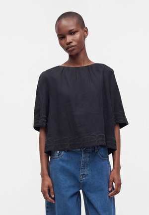 Donna con capelli corti che indossa una camicetta nera larga con orlo ricamato e maniche larghe, abbinata a jeans di denim blu.
