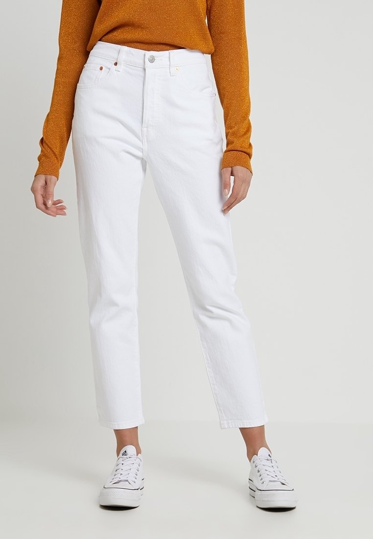 Personne portant un jean blanc taille haute droit, un pull orange à manches longues et des baskets blanches, debout devant un fond uni.
