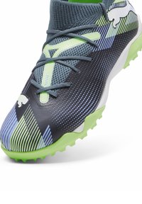 Zapatilla deportiva negra y verde con un upper texturizado, que presenta patrones a rayas en azul y verde, y una suela con tacos pronunciados para mayor tracción.