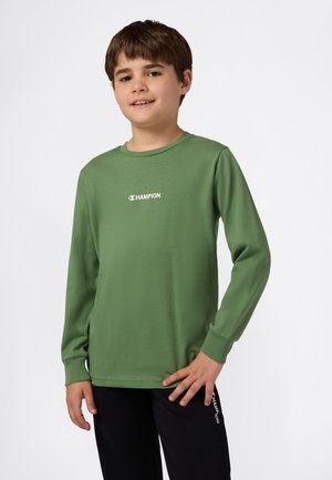Jongen in een groene long-sleeve Champion shirt en zwarte broek met Champion logo, staand met hand op de heup tegen een witte achtergrond.