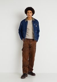 Jachetă din denim de culoare albastru închis, cu guler deschis, pulover bej dedesubt și pantaloni maro, largi, cu buzunare tip cargo. Pantofi maroon.