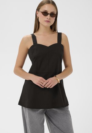 Femme portant un haut sans manches noir, un pantalon gris, des lunettes de soleil et un bracelet en or, se tenant les mains jointes devant un fond clair.