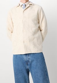 Krämfärgad texturerad skjorta med blommönster, med krage och knappar, bärs över blå denimjeans.