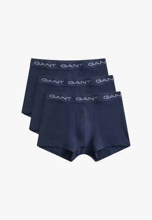Homme torse nu aux cheveux bouclés, debout contre un mur blanc, portant un boxer bleu marine avec la ceinture portant le logo GANT.