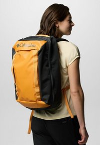 Schwarzer und orangefarbener Columbia-Rucksack mit gepolsterten Trägern, Reißverschlusstaschen und einer strukturierten Oberfläche. Auffälliges Logo auf der Vorderseite.