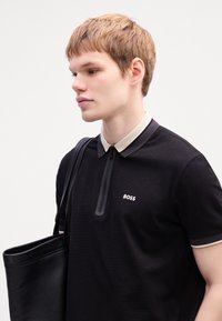 Juodas polo marškinėliai su dviejų tonų apykakle, užtrauktuku ir kontrastingomis pilkomis detalėmis. Yra balta "BOSS" logotipas. Juoda odinė rankinė.