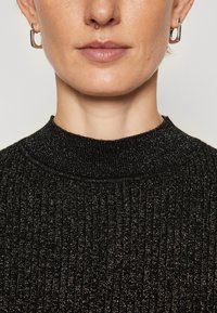 Svart ribbad tröja med hög krage, med en texturerad metallfinish. Silverfärgade ringörhängen framhäver stilen.