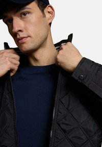 Czarna pikowana kurtka z zapięciem na zamek z przodu i paskami wewnętrznej podszewki, noszona na granatowej koszulce z okrągłym dekoltem. Zbliżenie na detale zamka i kołnierza.