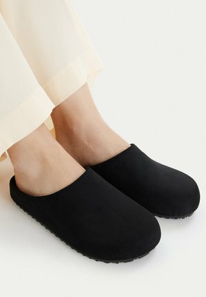 Füße, die einfache schwarze Slip-On-Clogs mit abgerundeten Zehen und strukturierten Sohlen tragen, kombiniert mit hellen Hosen.