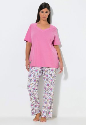 Femme debout pieds nus portant un haut rose ample à manches courtes et un pantalon de pyjama blanc avec des motifs abstraits colorés.