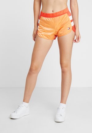 Žena v lesklých oranžových sportovních šortkách s bílými pruhy na stranách a bílými teniskami, stojící před jednoduchým světlým pozadím.