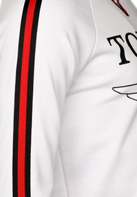 Vit långärmad skjorta med en svart logotyp och svarta/röda ränder längs ärmarna; slät tygtextur, närbild av designen.