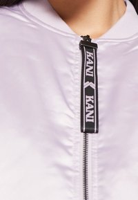 Lila bomberjacka med en glänsande textur, med en svart dragkedja och märkesdragretusch, plus ett ribbat krage och manschettdetaljer.