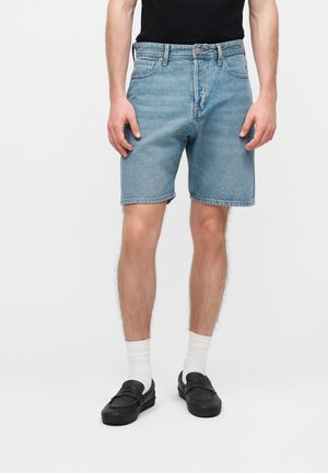 Muž nosi svetlomodré rifľové šortky, biele ponožky, čierne slip-on topánky a čiernu košeľu, stojí pred jednoliatym pozadím.