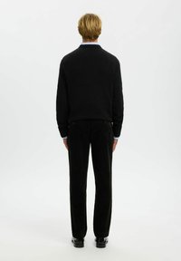 Blond mees seisab seljaga, seljas must pika varrukaga sviiter, mustad püksid, sinised pluusiääred käeotstes nähtavad ja mustad kingad.