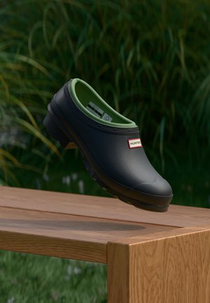 Μαύρο κλειστό παπούτσι Hunter slip-on με πράσινη εσωτερική επένδυση που αιωρείται πάνω από ένα ξύλινο παγκάκι, με ψηλό πράσινο χορτάρι στο θολό φόντο.