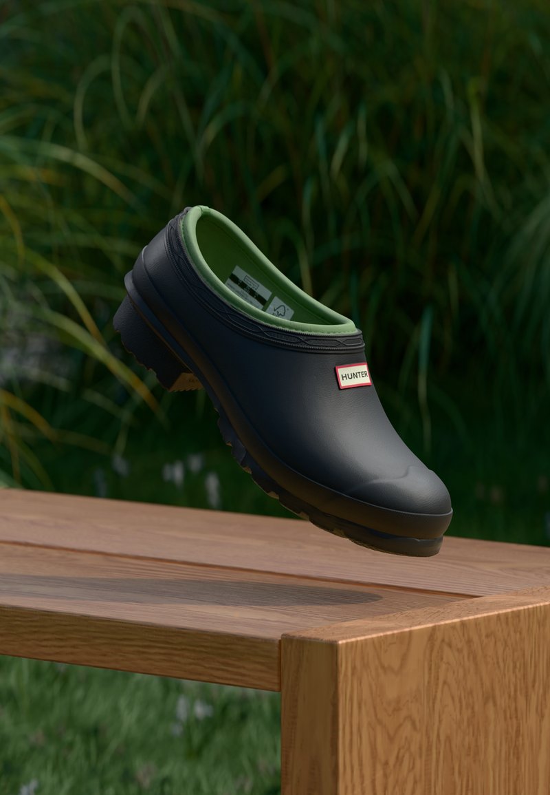 Μαύρο κλειστό παπούτσι Hunter slip-on με πράσινη εσωτερική επένδυση που αιωρείται πάνω από ένα ξύλινο παγκάκι, με ψηλό πράσινο χορτάρι στο θολό φόντο.