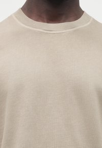 Světle béžové tričko s dlouhým rukávem a kulatým výstřihem, vyrobené z jemné látky s hladkou texturou a jemnými žebrovanými detaily u výstřihu.