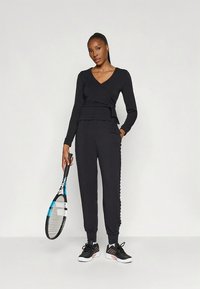 Fekete, hosszú ujjú bekötős felső és hozzáillő melegítőnadrág. Texturált anyag, testhez simuló kialakítás, fekete sportcipőkkel és teniszütővel párosítva.