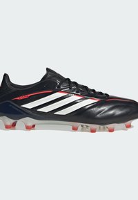 Chaussure de football noire avec une tige en cuir lisse et des accents rouges. Trois bandes blanches le long du côté, semelle extérieure transparente avec des crampons rouges.