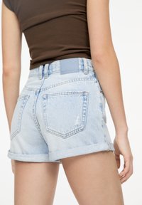 Svijetloplavi traper šortsevi s prevrnutim rubovima, s leđnim džepovima i sivom etiketom na pojasu. Oštećeni detalji oko džepova.