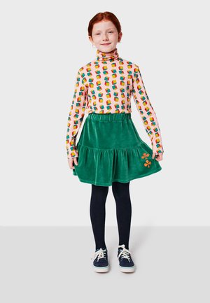 Long-sleeve, patterned top with colorful blocks; green velvet skirt with ruffle detail and orange floral embroidery; paired with navy sneakers.